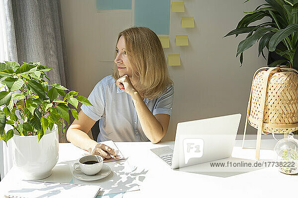 Lächelnder blonder Freiberufler sitzt mit Laptop am Schreibtisch im Heimbüro