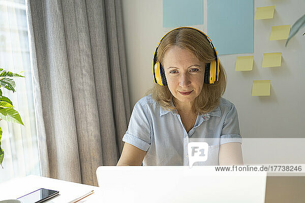 Mature businesswoman wearing wireless headphones using laptop working in home office