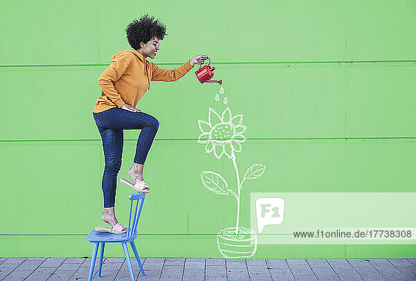 Woman standing on chair watering flowers painted on wall