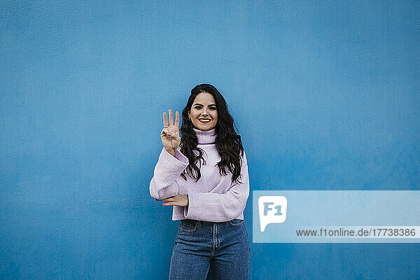 Lächelnde junge schöne Frau zeigt Nummer 3 vor blauer Wand