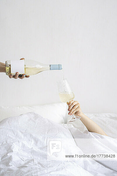 Hand pouring wine in wineglass held on bed at home