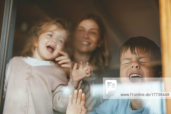 Playful boy pressing face on window glass by mother and sister