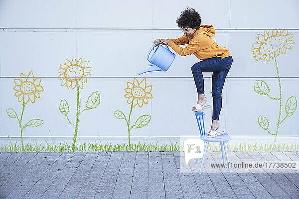 Woman standing on chair watering flowers painted on wall