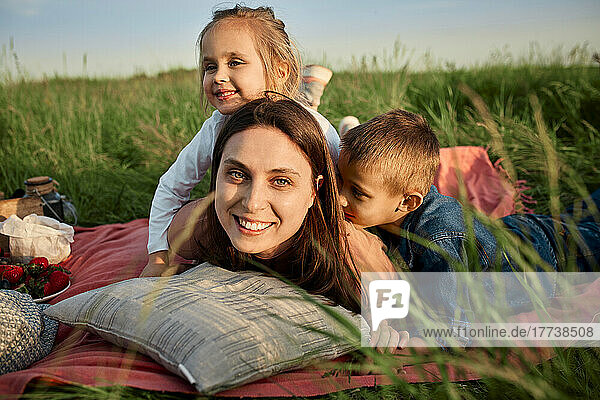 Smiling mother lying by children in picnic blanket at field