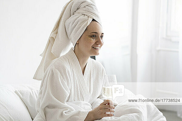 Happy woman wearing towel sitting on bed with wineglass at home