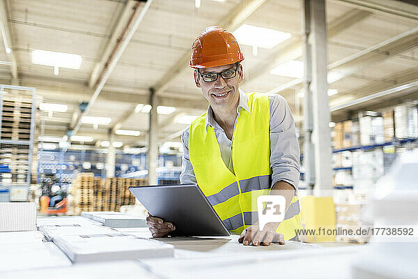 Smiling worker with tablet PC leaning on box in warehouse