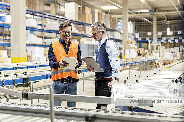 Worker discussing over tablet PC with manager standing by conveyor belt in warehouse