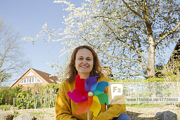 Happy woman with pinwheel toy sitting in front of tree at playground