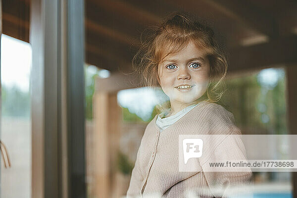 Lächelndes süßes Mädchen zu Hause durch Fensterglas gesehen