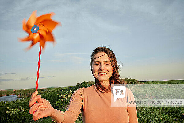 Smiling young woman holding pinwheel toy with eyes closed on field at sunset