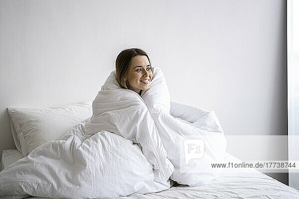 Glückliche Frau  in eine Decke gehüllt  sitzt zu Hause im Bett
