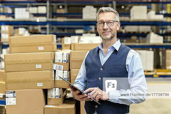 Smiling manager wearing eyeglasses standing with tablet PC in front of cardboard boxes at warehouse
