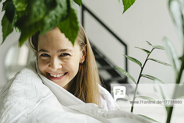 Smiling woman wrapped in blanket by plants at home