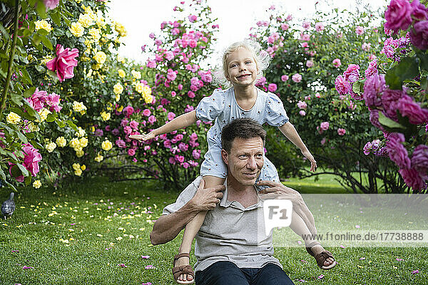Playful father carrying daughter on shoulders in front of flowering plants at garden