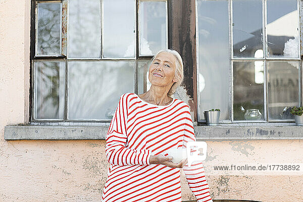 Smiling woman holding cup standing in front of house