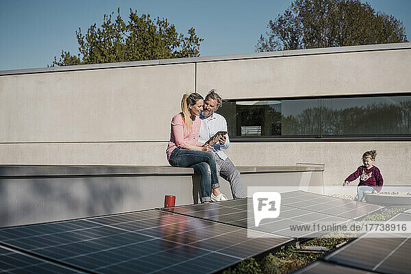 Woman holding tablet PC sitting by man with daughter playing by solar panel on sunny day