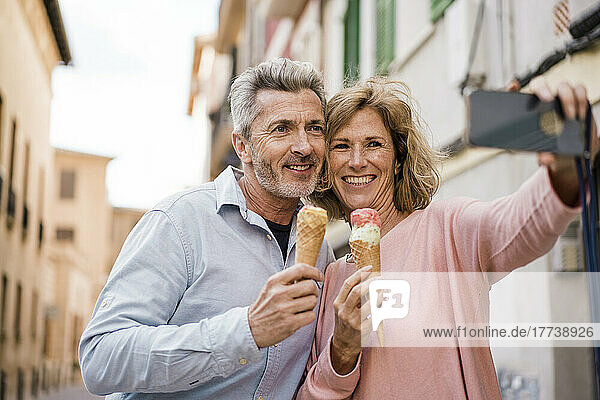 Smiling couple with ice cream taking selfie on mobile phone in city