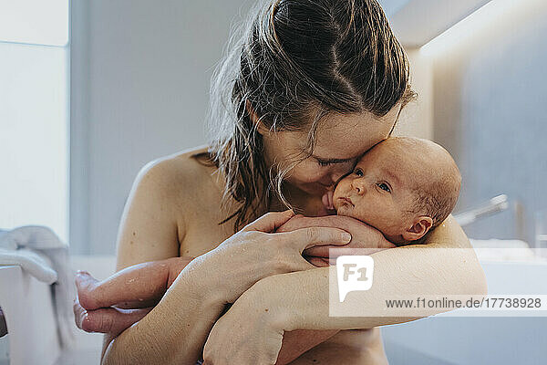 Naked mother cuddling cute baby in bathtub at home