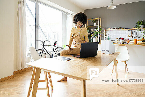 Smiling woman holding mobile phone looking at laptop sitting on table