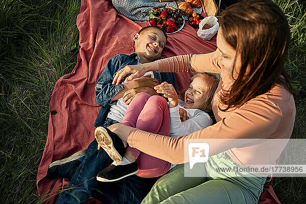 Mother tickling children lying on picnic blanket in field