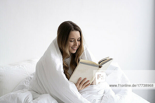 Smiling young woman wrapped in white blanket reading book on bed at home