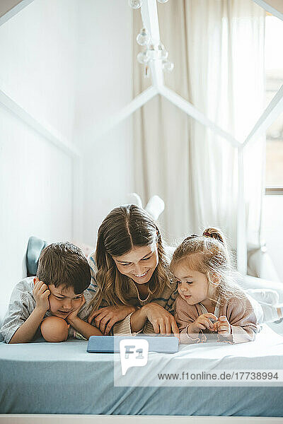 Smiling woman with son and daughter looking at tablet PC lying on bed
