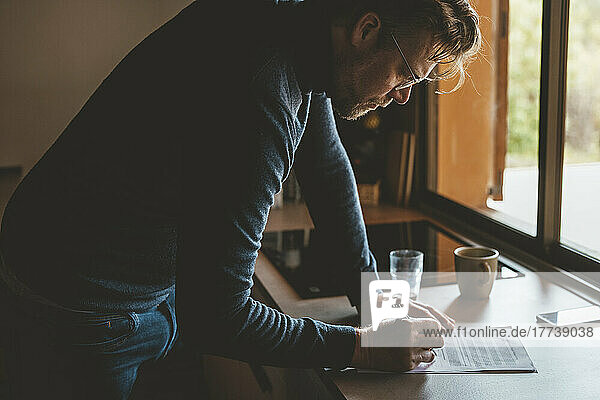 Mature man leaning on kitchen counter examining document at home