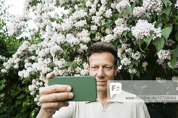 Smiling man taking selfie through smart phone in front of flowering plant