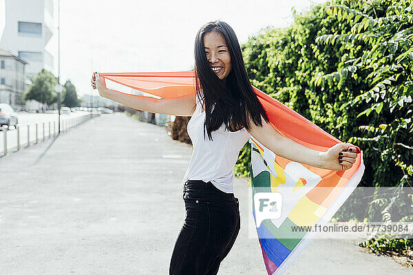 Happy woman with arms outstretched standing with rainbow flag on sunny day