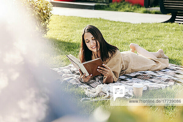 Smiling young woman reading book lying on picnic blanket at park