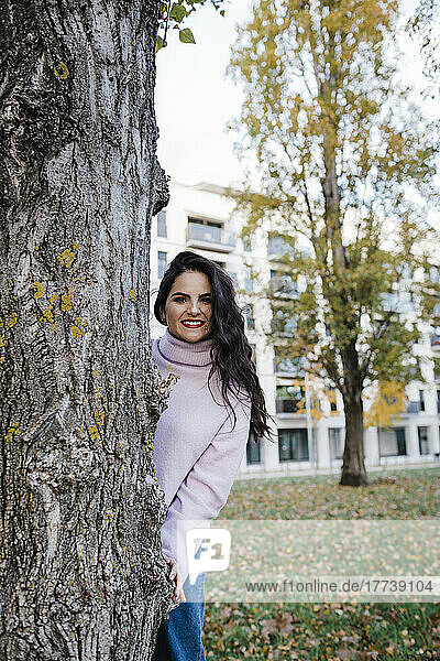 Smiling young beautiful woman enjoying autumn standing behind tree trunk at park