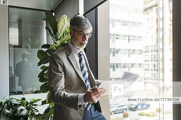 Businessman with documents using tablet PC standing by window in office