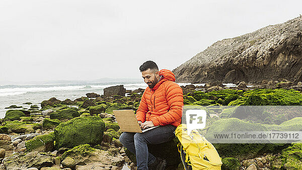 Smiling man using laptop sitting with backpack on rock