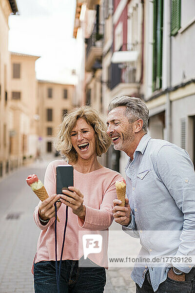 Cheerful mature couple with ice cream taking selfie on smart phone in city