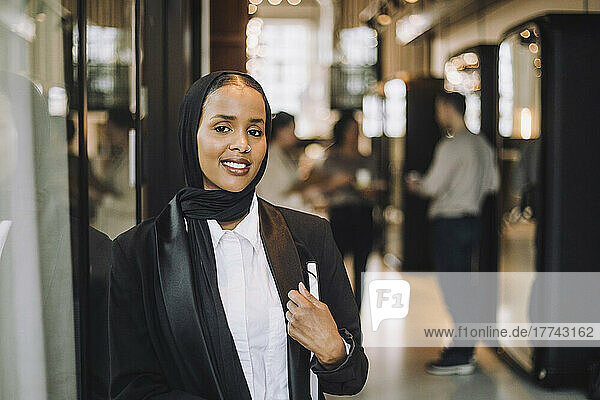 Lächelnde junge Unternehmerin mit Kopftuch  die ein Tagebuch hält  während sie im Büro steht