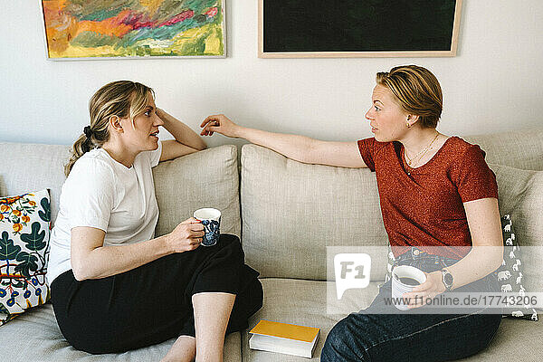 Lesbian couple talking while having coffee on sofa in living room at home