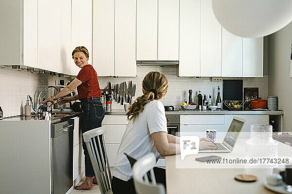 Freelancer working on laptop while talking with girlfriend doing chores in kitchen at home