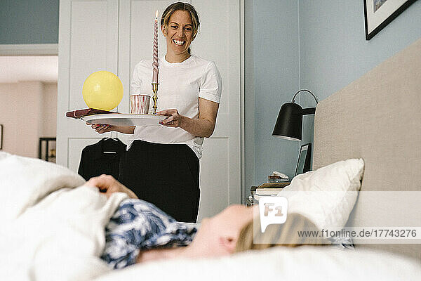 Smiling woman with candle and surprise birthday gift standing in front of girlfriend lying on bed at home