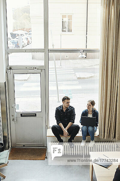 Junge Geschäftskollegen diskutieren miteinander  während sie vor einem Glasfenster im Büro sitzen