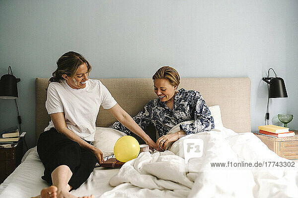 Smiling lesbian couple looking at birthday surprise on bed in bedroom