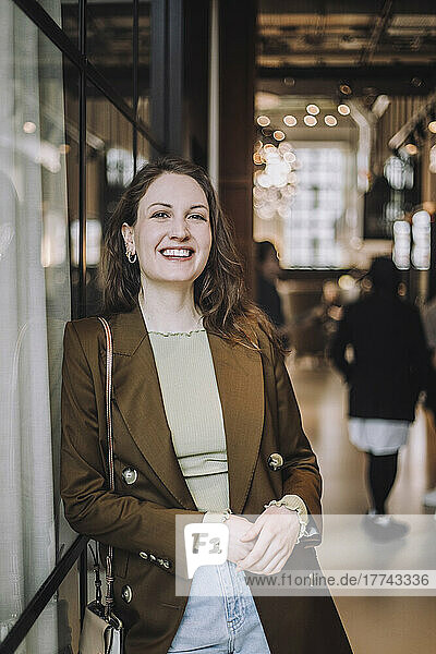 Portrait of happy mid adult businesswoman leaning on wall while standing in creative office