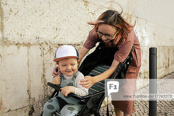 Lächelndes männliches Kleinkind im Kinderwagen mit Mutter auf dem Fußweg