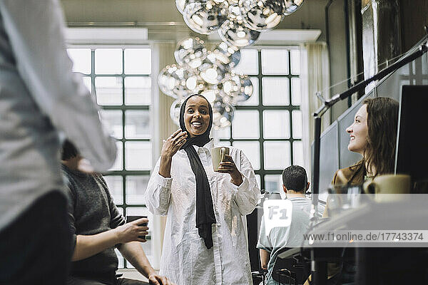 Lächelnde junge Geschäftsfrau  die eine Kaffeetasse hält  während sie mit Kollegen in einem kreativen Büro Ideen austauscht