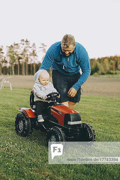 Father giving ride to son sitting in toy tractor at farm