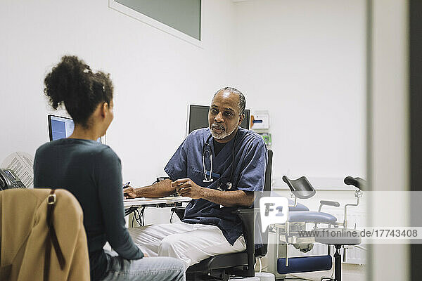 Reifer männlicher Arzt im Gespräch mit einer Patientin  die am Schreibtisch in einer medizinischen Klinik sitzt