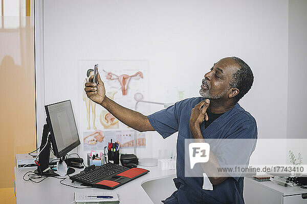 Male otolaryngologist doing online consultation through video call on smart phone while sitting at desk in clinic