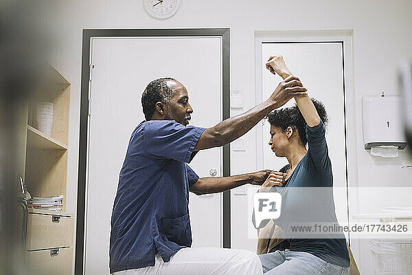 Side view of male doctor giving physical therapy to female patient sitting with hand raised in hospital