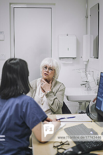 Ältere Patientin erklärt dem Arzt am Schreibtisch sitzend in einer medizinischen Klinik ihr Halsproblem