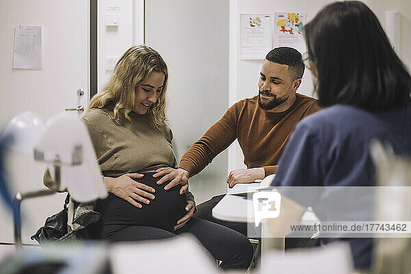 Smiling man touching stomach of pregnant woman while discussing with gynecologist in medical clinic