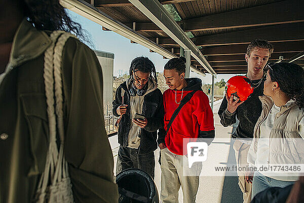 Young man sharing smart phone with male friend while walking on railroad station platform